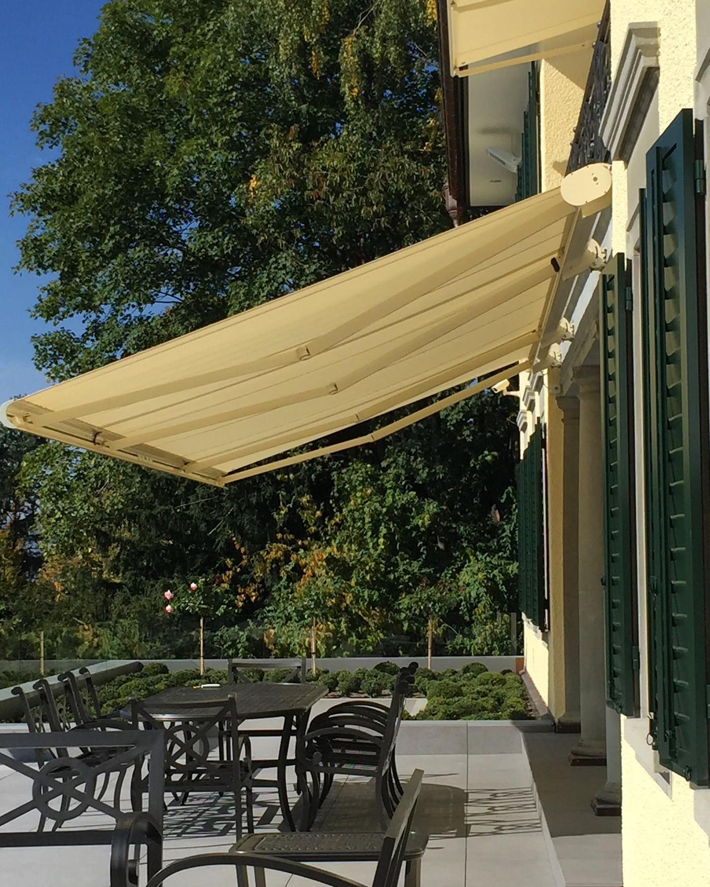Cremefarbene Gelenkarmmarkise auf einer Villenterrasse mit grünen Klappläden